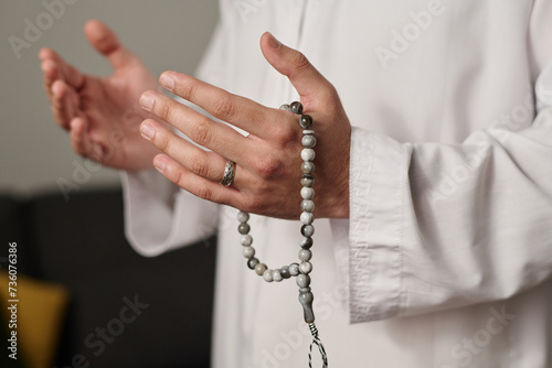 Hands of unrecognizable man in white thwab holding rosary beads during silent pray or while worshipping Allah on religious occasion