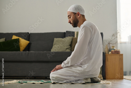 Young Muslim man in white skullcap and thobe standing on knees on rug and performing namaz prayer against couch with cushions photo
