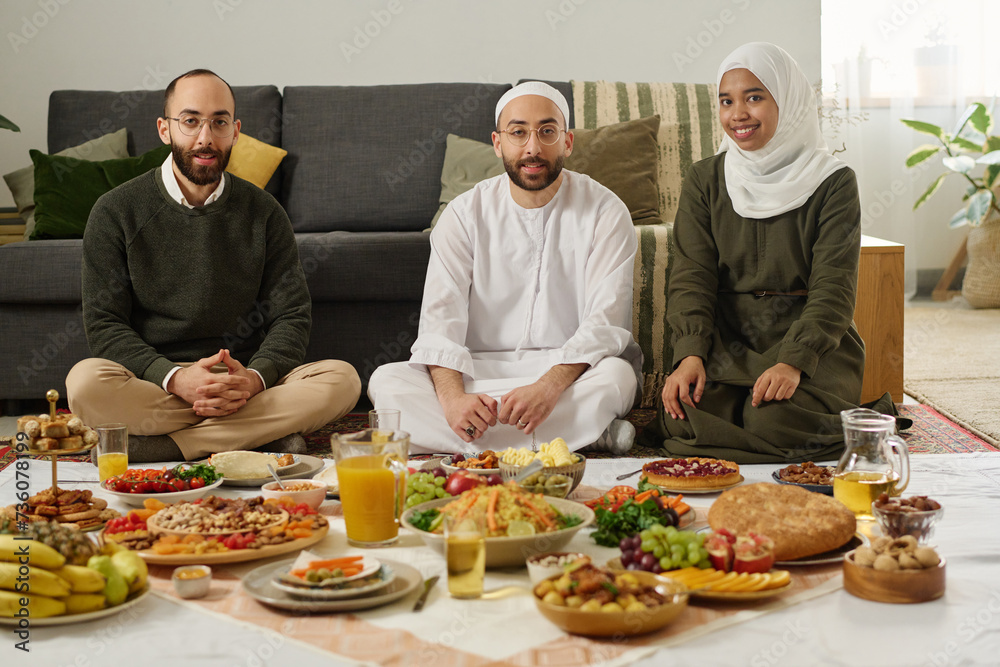 Happy young Muslim couple in traditional clothes and their guest sitting on the floor with homemade food on tablecloth during dinner Happy young Muslim couple in traditional clothes and their guest sitting on the floor with homemade food on tablecloth during dinner