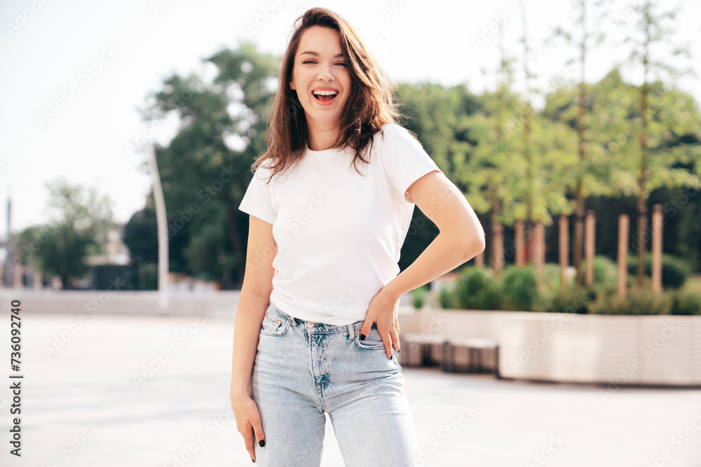 Portrait of young beautiful smiling hipster woman in trendy summer white t-shirt and jeans clothes. Sexy carefree model posing on the street background at sunset. Positive model outdoors