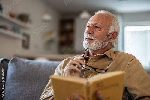 Home, relax and senior man with a book, retirement and calm with a story in a living room. Old person, calm and pensioner in a lounge, novel or literature with a hobby, self help or reading on a sofa. photo