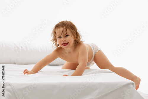 Radiant and playful child  little baby-girl in diaper enjoying fun time on comfortable white bedspread against white background. Concept of childhood  motherhood  life  birth. Copy space for ad