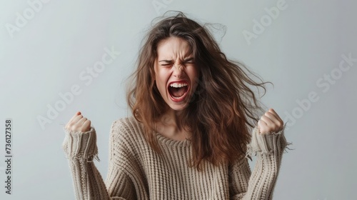 A young woman in a beige sweater passionately screams, fists clenched, eyes closed, and hair disheveled, expressing intense emotion against a neutral background
