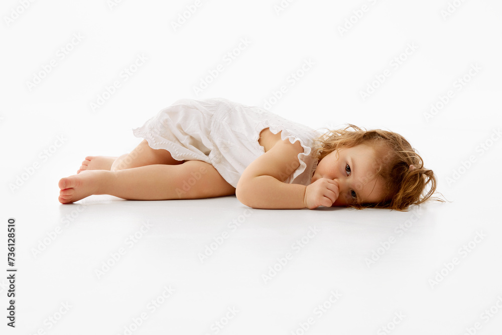 Cute little baby with curly and wet hair in frilly dress resting on floor with thoughtful expression against white background. Concept of childhood, carefree, motherhood, life, birth. Copy space. Ad Cute little baby with curly and wet hair in frilly dress resting on floor with thoughtful expression against white background. Concept of childhood, carefree, motherhood, life, birth. Copy space. Ad