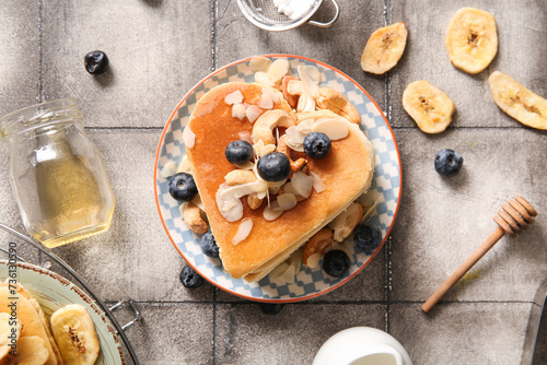 Plate with tasty pancakes in shape of heart, dried banana, blueberry and honey on grey tile background. Valentine's Day celebration