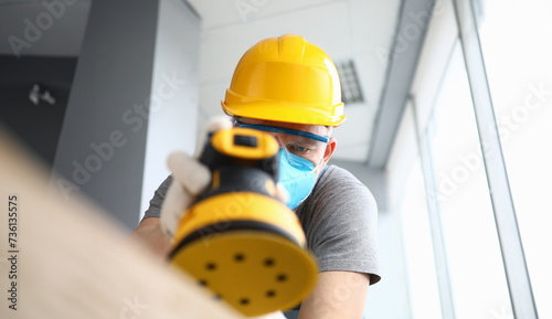 Close-up of construction worker using sander machine wearing protective yellow helmet and gloves. Repairman polishing wood in apartment. Renovation concept