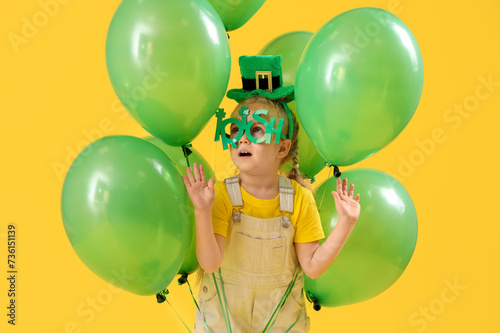 Cute little girl with leprechaun's hat, novelty glasses and balloons on yellow background. St. Patrick's Day celebration