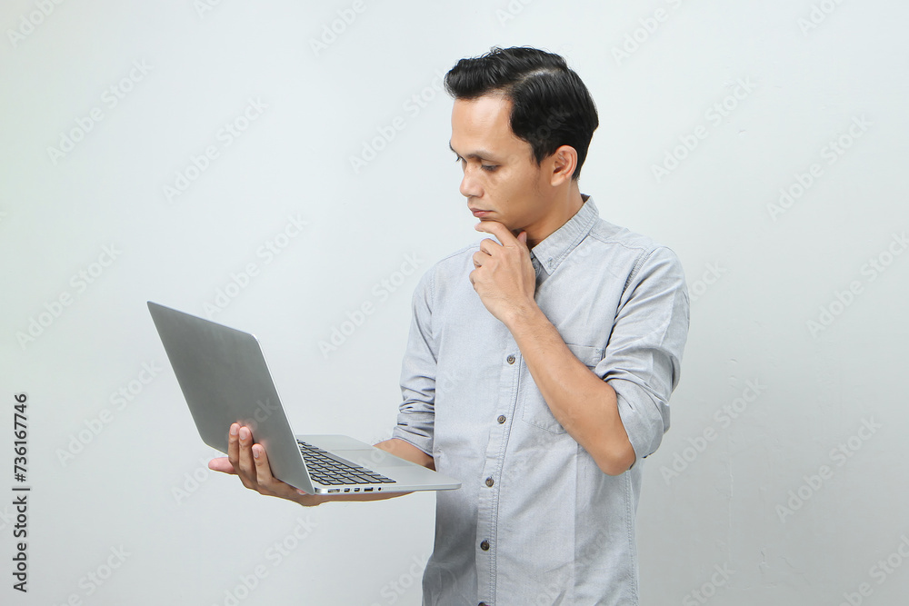 Pensive young Asian Indonesian man holding laptop computer with a serious face thinking about a question, thoughtful about confusing idea on isolated background