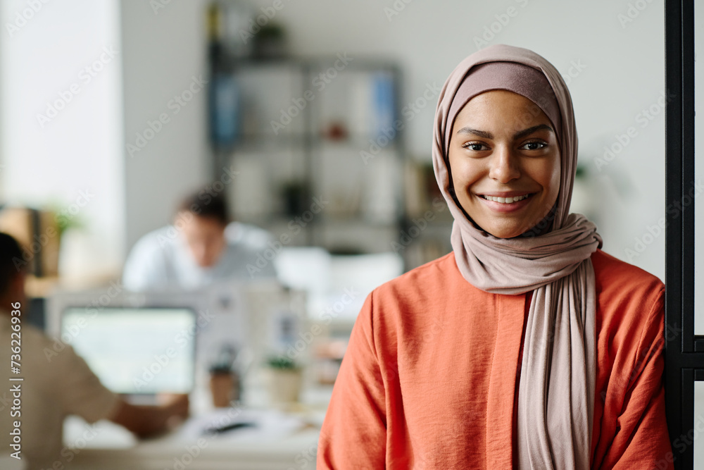 Happy young Muslim female entrepreneur or manager in hijab looking at camera with smile while standing in coworking space