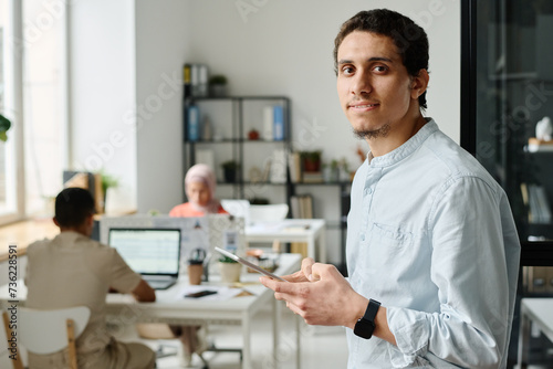 Young Muslim male entrepreneur or office manager with tablet looking at camera with smile while standing against colleagues in coworking space