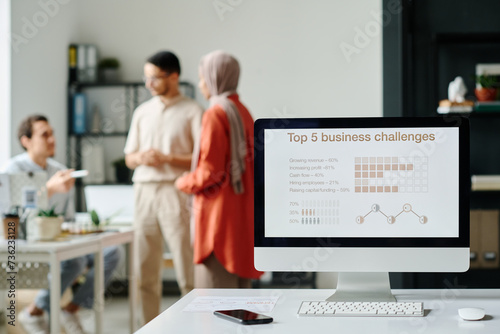 Screen of desktop computer with graphic data standing on desk against group of modern Muslim brokers having meeting in office photo