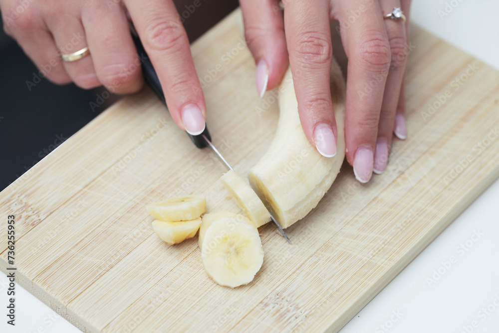 Woman hands cutting banana. Banana slices. Brown wooden cutting board. Female fingers holding knife. Bananas salad perparation. Healthy breakfast meal.