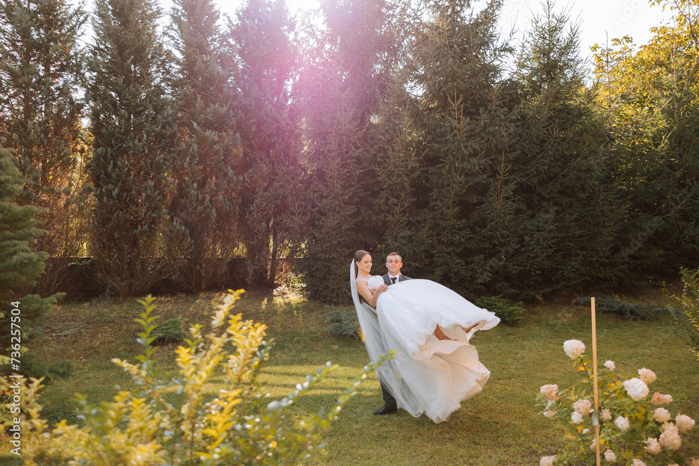 Stylish groom holds beautiful bride in long white dress in his arms and dances on green grass in autumn park, forest under sunlight. Wedding photography, portrait of smiling newlyweds. Stylish groom holds beautiful bride in long white dress in his arms and dances on green grass in autumn park, forest under sunlight. Wedding photography, portrait of smiling newlyweds.