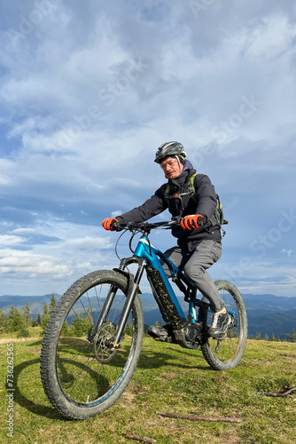 Cyclist man riding electric mountain bike outdoors. Male tourist biking along grassy trail in the mountains, wearing helmet and backpack. Concept of sport, active leisure and nature.