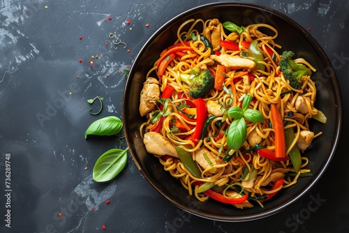 Top view of stir fried noodles with veggies and chicken in a black bowl on a dark background with space for text
