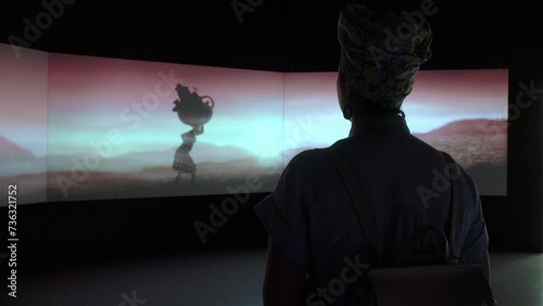 A girl with a backpack standing in front of a large, curved screen in a museum. The screen displays an installation on the theme of “the difficult path of a woman in Africa”.
