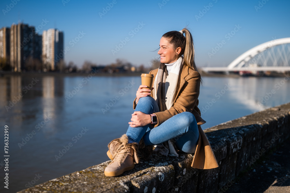 Beautiful woman in warm clothing enjoys drinking coffee and resting by the river on a sunny winter day.	