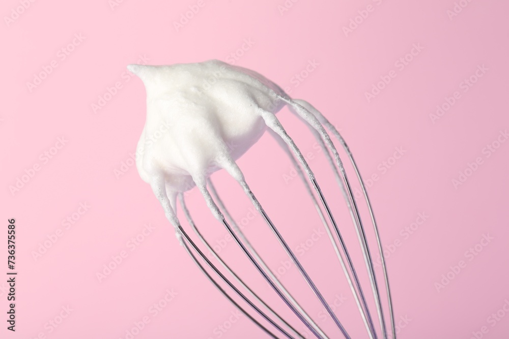 Whisk with whipped egg whites on pink background, closeup