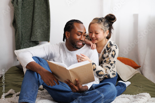 Smiling young asian woman hugging african american boyfriend with book on bed at home