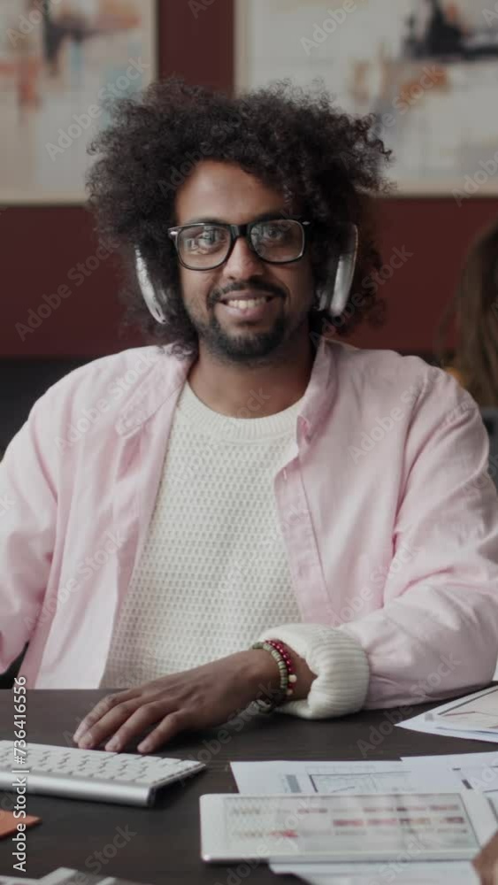 Vertical Waist up slowmo portrait of Biracial male interior designer in wireless headphones sitting at desk with blueprints on it and smiling at camera, working from home officeVertical Waist up slowm Vertical Waist up slowmo portrait of Biracial male interior designer in wireless headphones sitting at desk with blueprints on it and smiling at camera, working from home officeVertical Waist up slowm