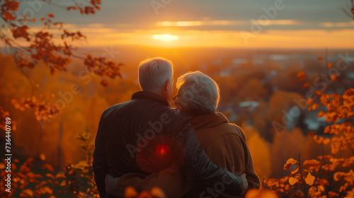 An elderly couple happily embracing while watching the sunset from the top of a mountain with a beautiful landscape. They represent health in old age