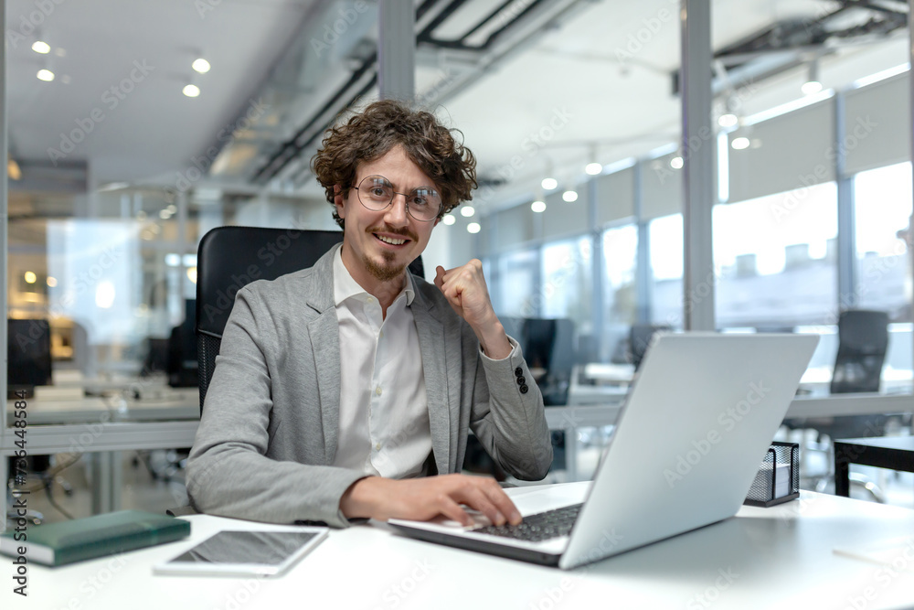 Smiling young businessman with curly hair focused on work at his laptop in a well-lit modern corporate office setting. Smiling young businessman with curly hair focused on work at his laptop in a well-lit modern corporate office setting.