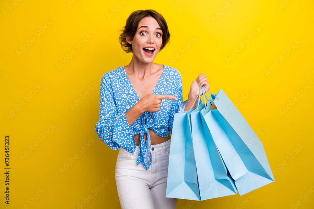 Portrait of ecstatic girl with short hairstyle wear blue blouse directing at shopping bags staring isolated on yellow color background Portrait of ecstatic girl with short hairstyle wear blue blouse directing at shopping bags staring isolated on yellow color background