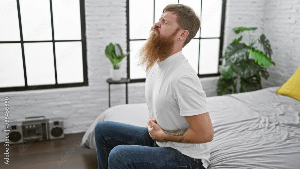 Tormented young adult redhead guy touching his aching stomach while sitting miserably on bed in bedroom, suffering from stomach pain, an indoor awakening ordeal Tormented young adult redhead guy touching his aching stomach while sitting miserably on bed in bedroom, suffering from stomach pain, an indoor awakening ordeal