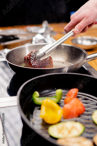 close-up of seared beef steak with rosemary in tongs on frying pan next to vegetables