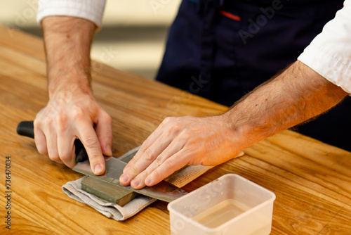 close-up of a chef in a white jacket in the kitchen at the table sharpens a knife on a green stove