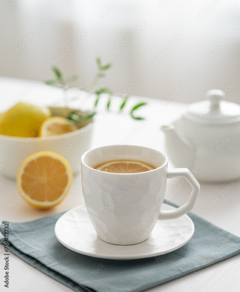 A cup of tea with lemon and a teapot on a white table against the background of a kitchen window close up. A cup of tea with lemon and a teapot on a white table against the background of a kitchen window close up.