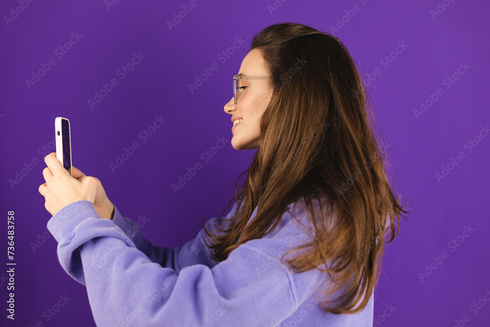 Great application or website. Portrait of excited woman using her mobile phone, isolated on purple studio background, standing side. Positive lady holding gadget, looking at phone, texting, messaging. Great application or website. Portrait of excited woman using her mobile phone, isolated on purple studio background, standing side. Positive lady holding gadget, looking at phone, texting, messaging.