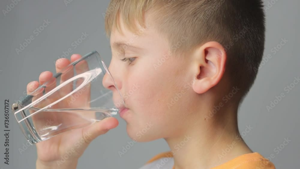Cute boy drinks clean water from a glass on a gray background. portrait of a child with a glas