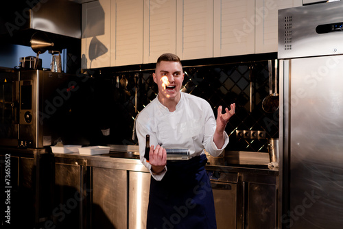 A chef in a white jacket in a professional kitchen plays with a working burner in his hand and smiles