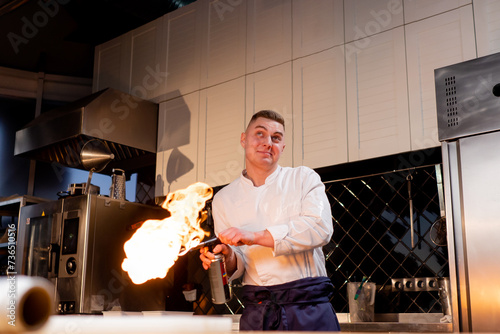 A chef in a white jacket in a professional kitchen plays with a working burner in his hand and smiles