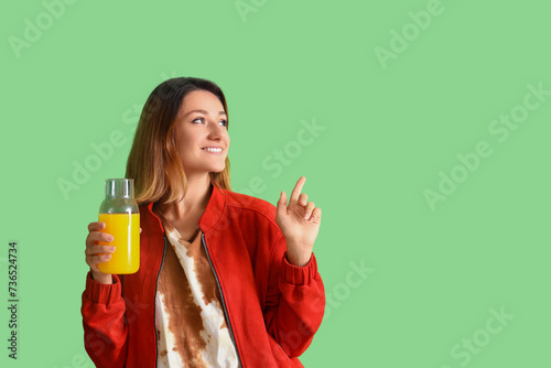 Young woman with glass bottle of juice pointing at something on green background