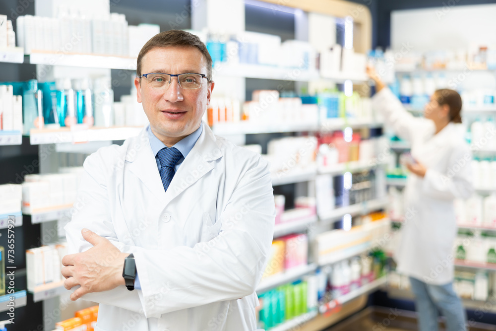 Smiling middle-aged man apothecary standing in front of shelves full of goods in big pharmacy