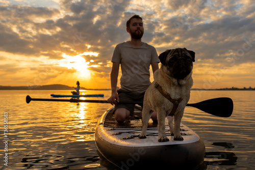 Man paddle boarding at lake during sunset together with pug dog. Concept of active tourism and supping with pets. Brave Dog Standing on SUP Board and enjoy lifestyle on summer petfriendly vacation photo