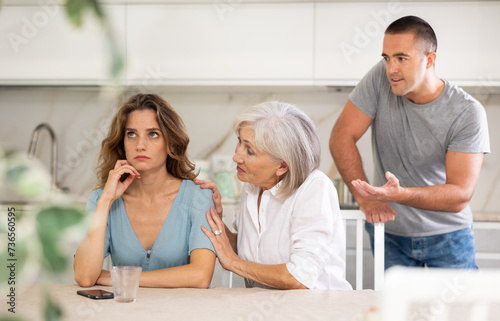Adult man and elderly woman during family quarrel with adult woman in kitchen