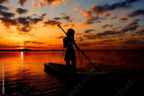 silhouette of woman on inflatable SUP board and paddling through shining water surface. aesthetically wide shot. Freedom happy female at sunset on a lake © Yekatseryna