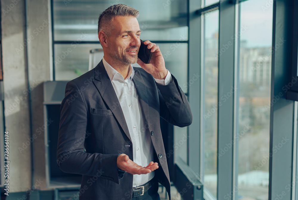 Smiling businessman is talking phone with client while standing in modern coworking near window Smiling businessman is talking phone with client while standing in modern coworking near window