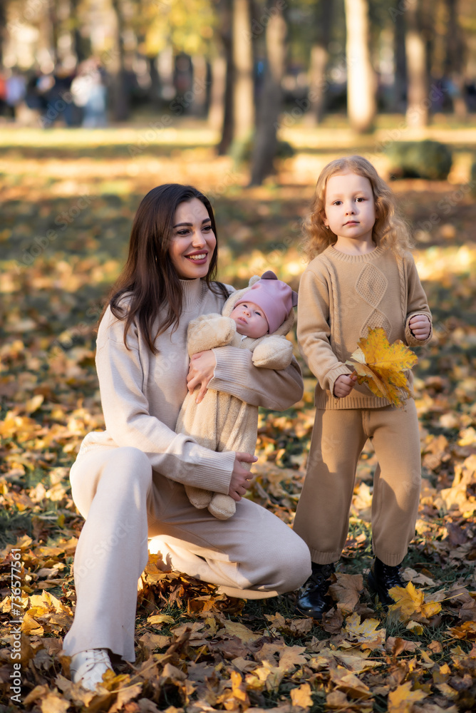 Young mother holds her youngest daughter in her arms in an autumn park. The eldest daughter stands nearby and holds leaves in her hands Young mother holds her youngest daughter in her arms in an autumn park. The eldest daughter stands nearby and holds leaves in her hands