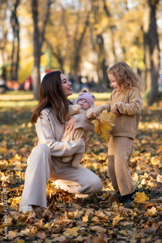 Young mother holds her youngest daughter in her arms in an autumn park. The eldest daughter stands nearby and holds leaves in her hands