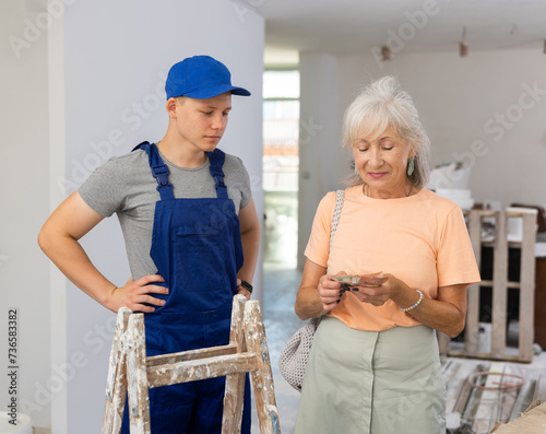Mature woman pays a fifteen-year-old worker in uniform for construction work performed in an apartment during renovation photo