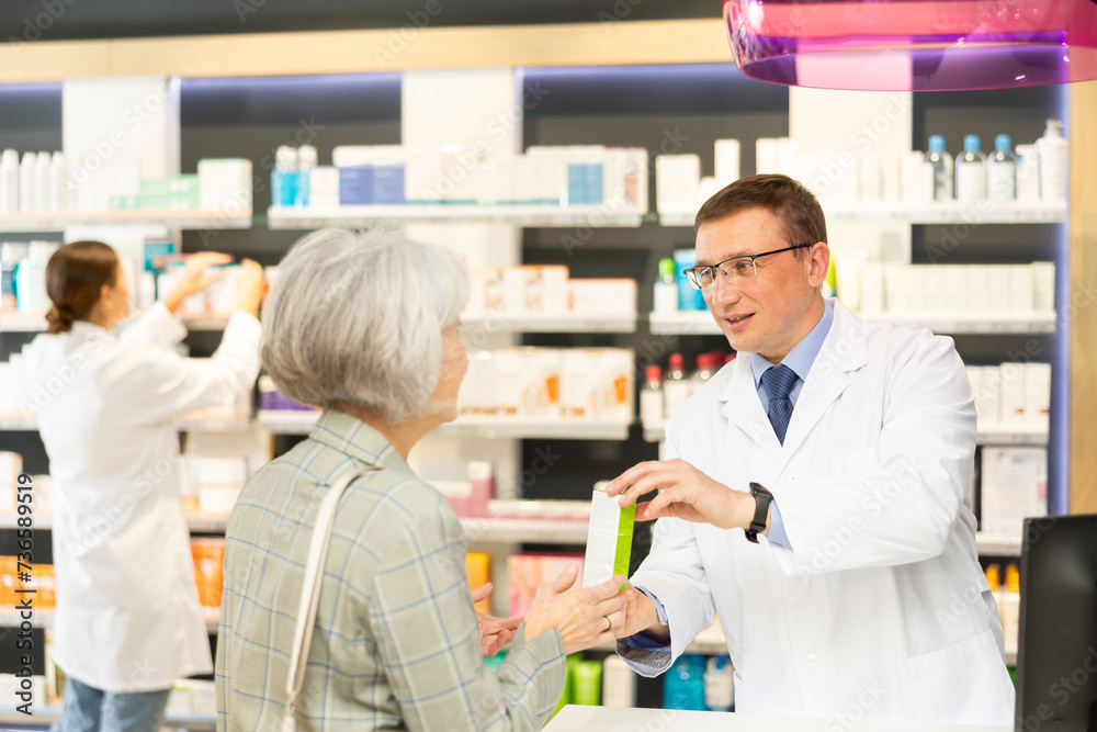 Polite middle-aged male pharmacist consulting old woman costumer about care product in box in chemist's shop