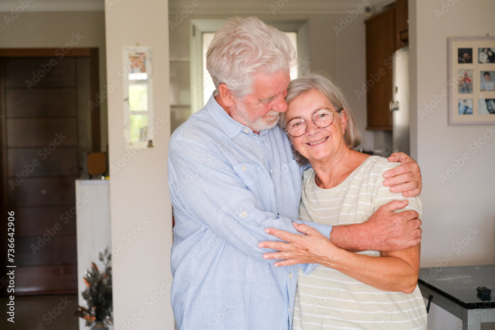 Senior couple hug and smile with apartment in background. Concept of life together forever elderly people. Mature man and woman enjoy indoor leisure activity with a portrait inside living room house Senior couple hug and smile with apartment in background. Concept of life together forever elderly people. Mature man and woman enjoy indoor leisure activity with a portrait inside living room house