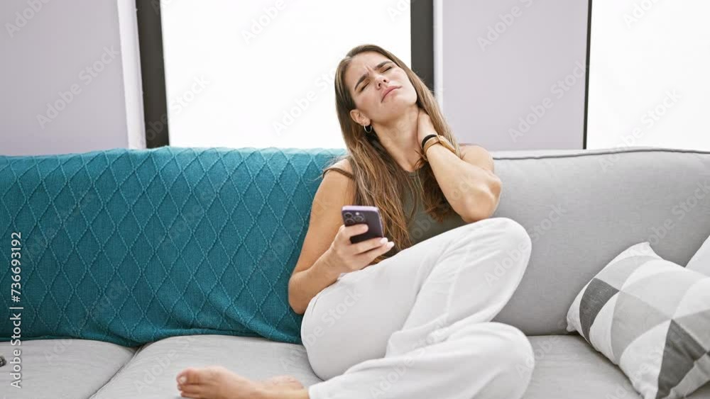 Stressed young beautiful hispanic woman, sitting at home on the sofa, suffers cervical pain from excessive smartphone usage Stressed young beautiful hispanic woman, sitting at home on the sofa, suffers cervical pain from excessive smartphone usage