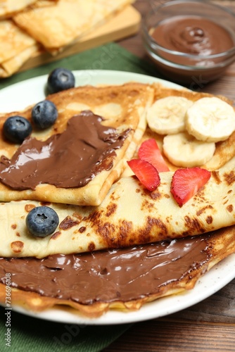Tasty crepes with chocolate paste, banana and berries on wooden table, closeup