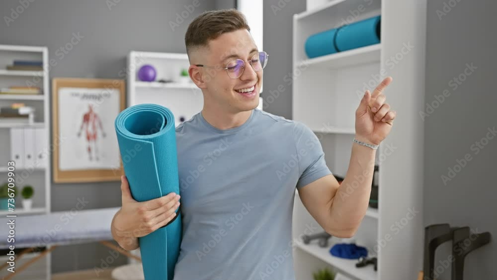 Optimistic young hispanic man at rehab clinic, smiling cheerfully, confidently pointing to the side with hand while holding yoga mat Optimistic young hispanic man at rehab clinic, smiling cheerfully, confidently pointing to the side with hand while holding yoga mat