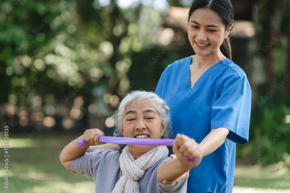 Fototapeta premium Compassionate Asian woman provides care to elderly person in wheelchair outdoors. Engaging in physical therapy, happiness, encouraging positive environment for mature individuals with grey hair.
